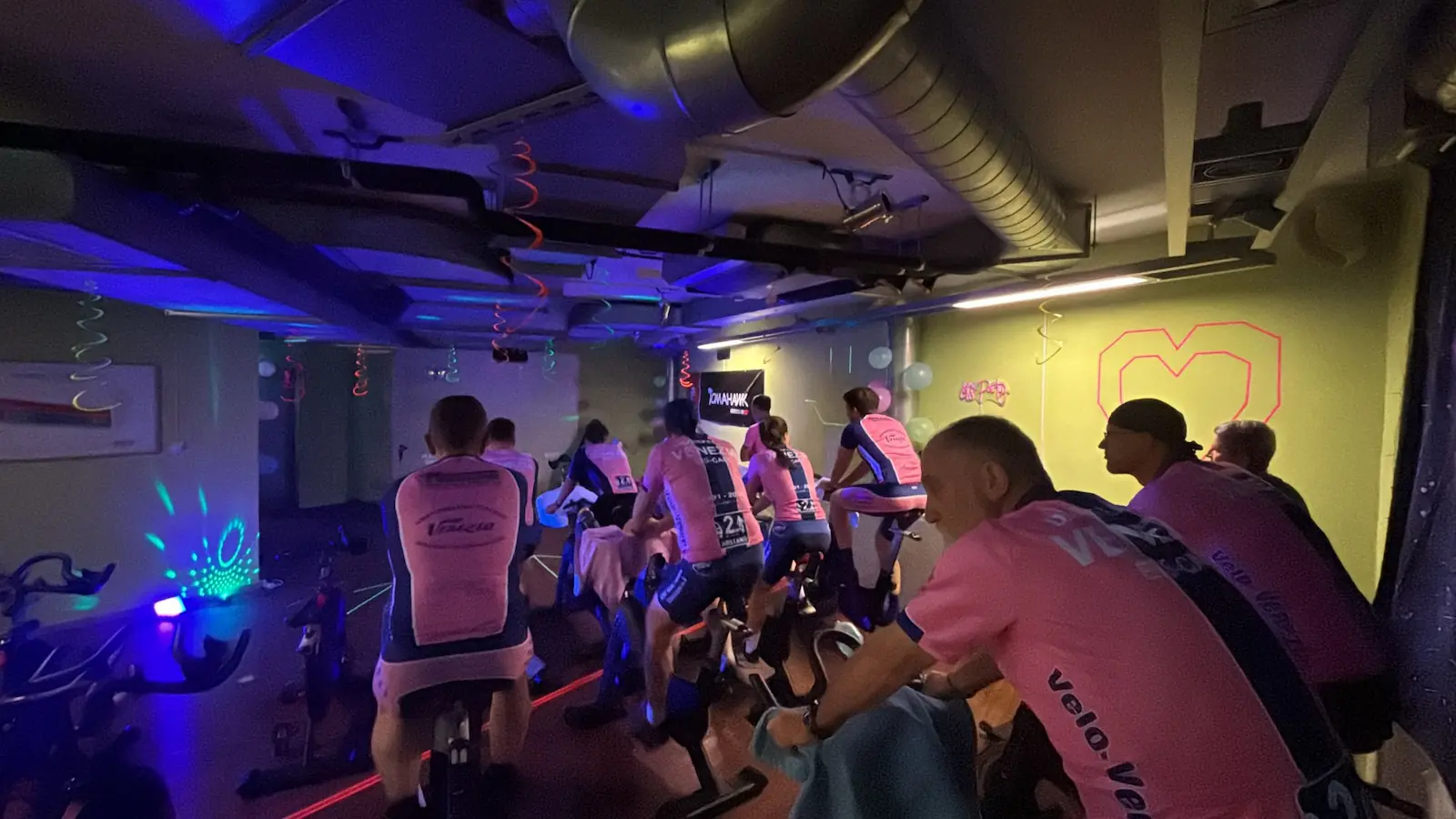 Spinning-Abend: die Gruppe in Vereinstrikots auf Indoor-Bikes bei farbigem Licht
